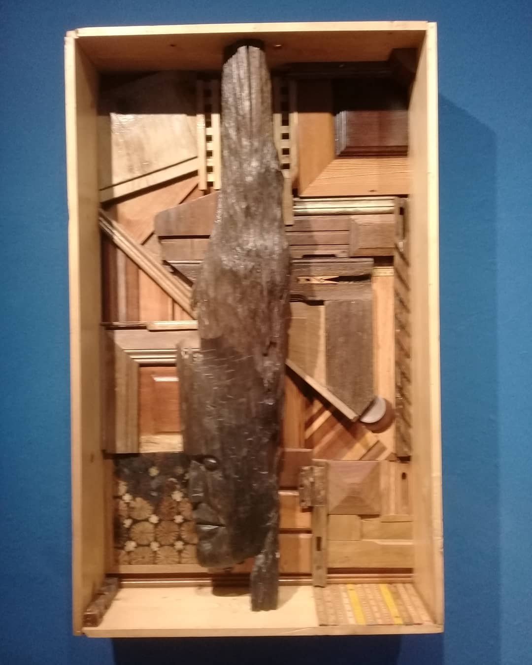 Caixa 06, relevo em madeira com uma forma vertical esculpida contra blocos geométricos em uma caixa clara sobre fundo azul.
