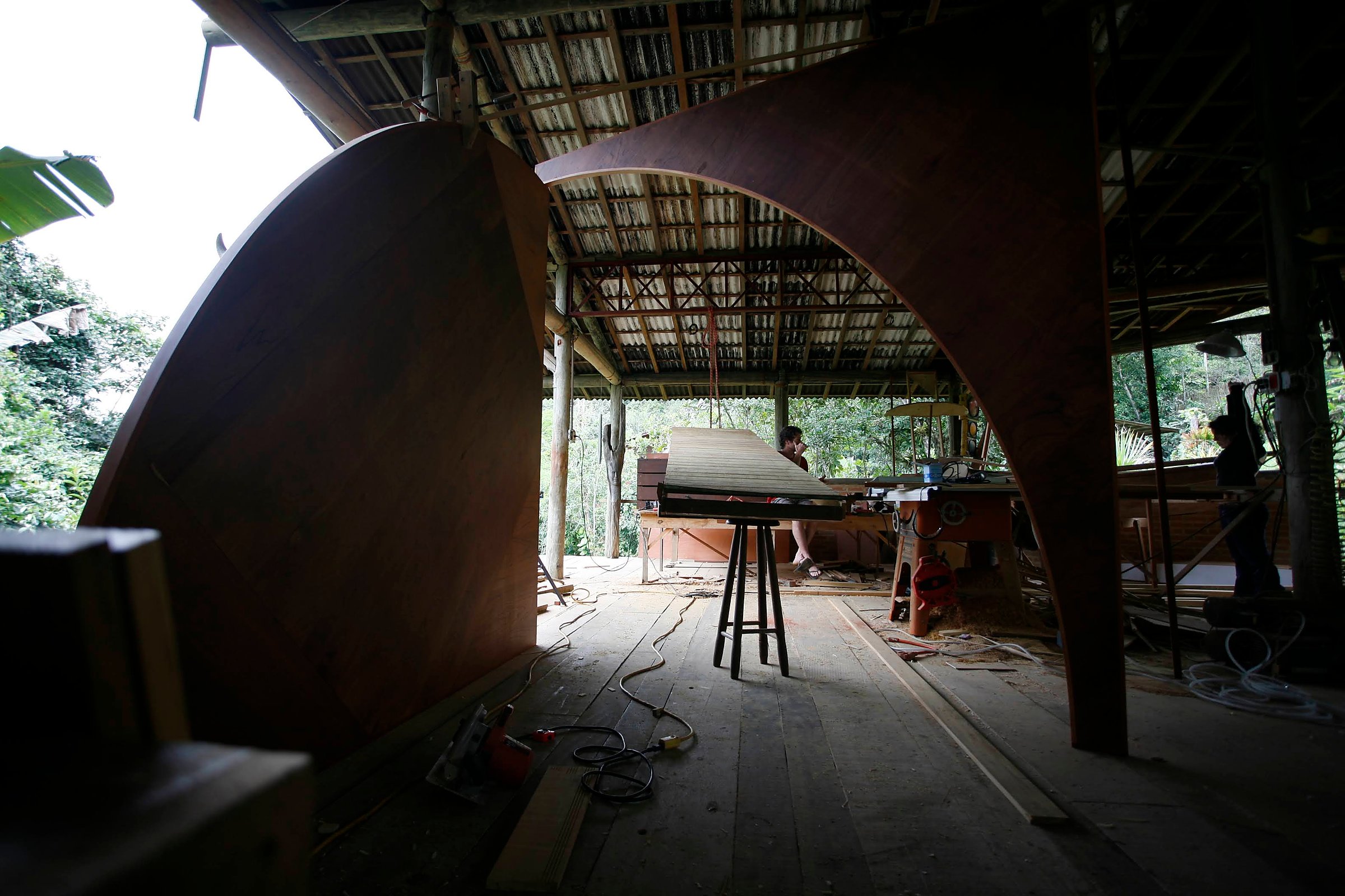 A escultura móvel Duo em compensado naval de cedro e muiracatiara ergue-se em uma oficina aberta com duas figuras ao fundo.