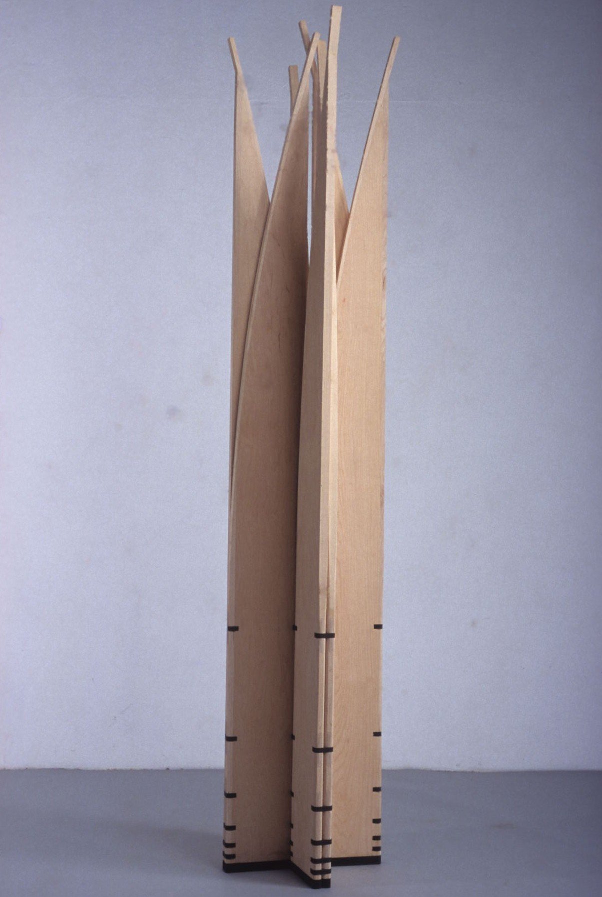 Mobile sculpture of pointed vertical wooden sections gathered into a closed upright column on a grey floor.