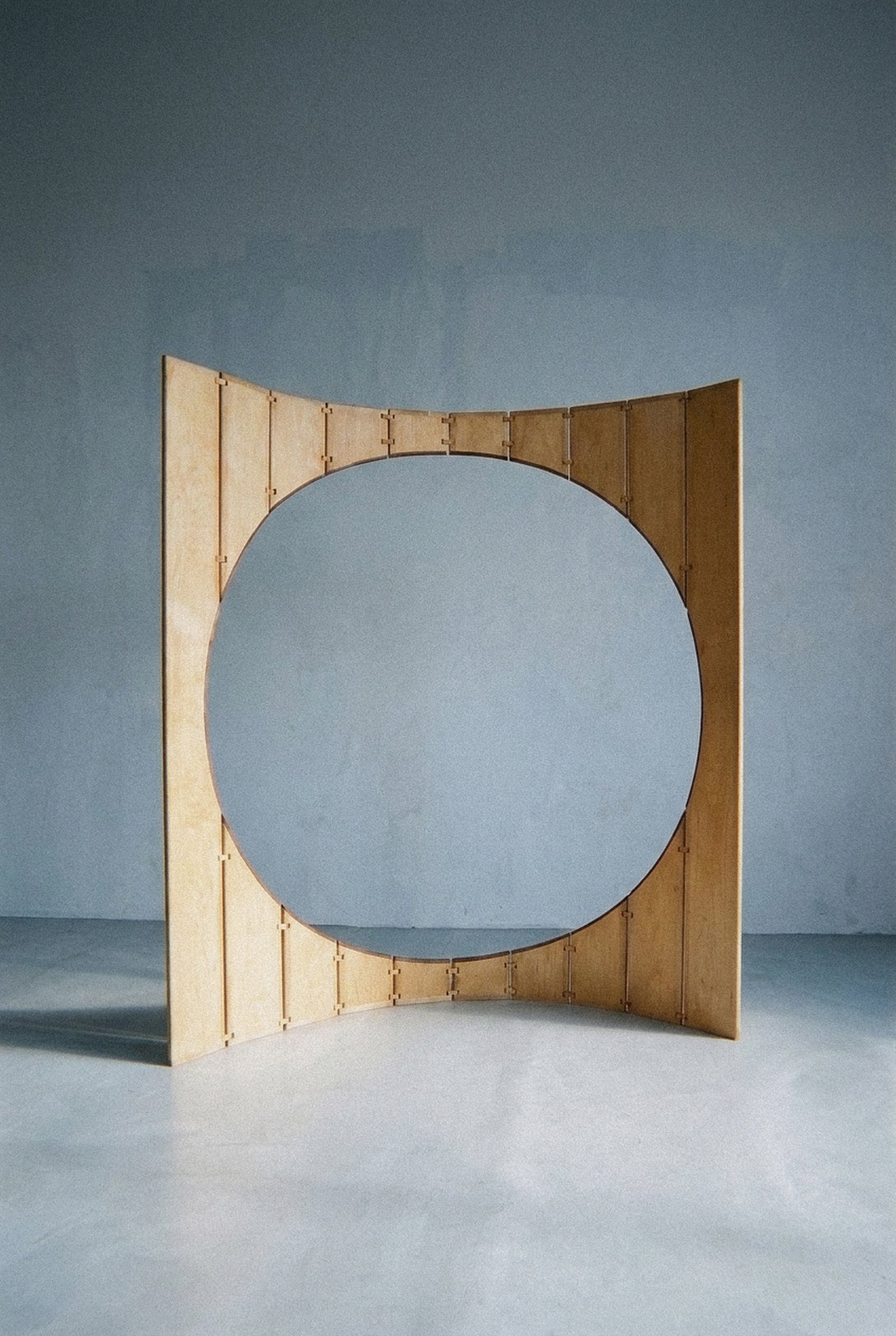 A escultura móvel Ó em madeira e latão forma um arco aberto com grande vão circular sobre piso cinza.