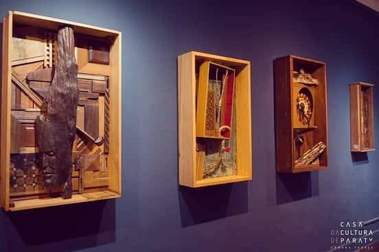 Quatro caixas de madeira com relevos internos fixadas em linha sobre uma parede azul escura em uma galeria.