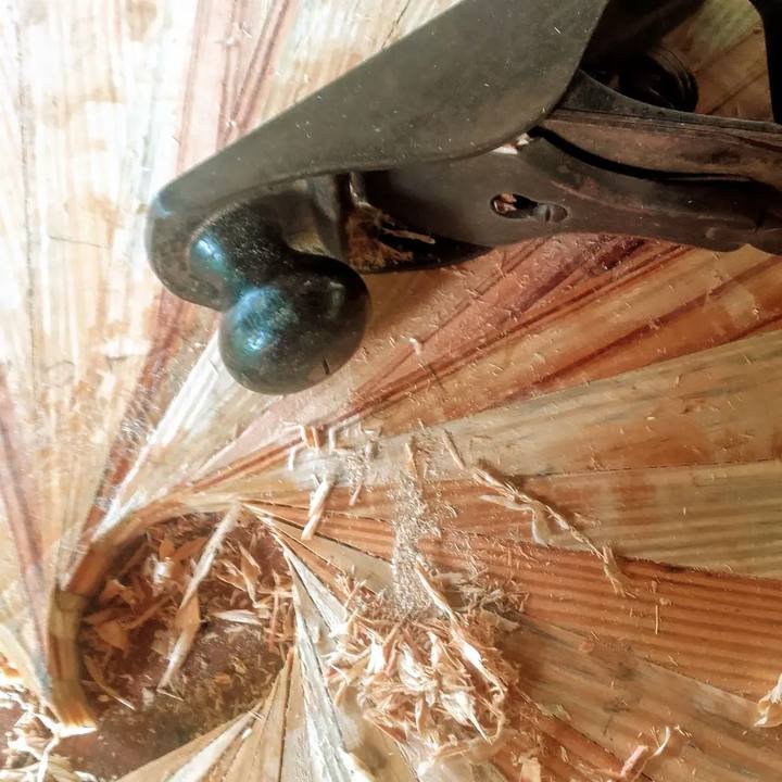 A hand plane shapes a radial wooden surface covered with fine wood shavings.